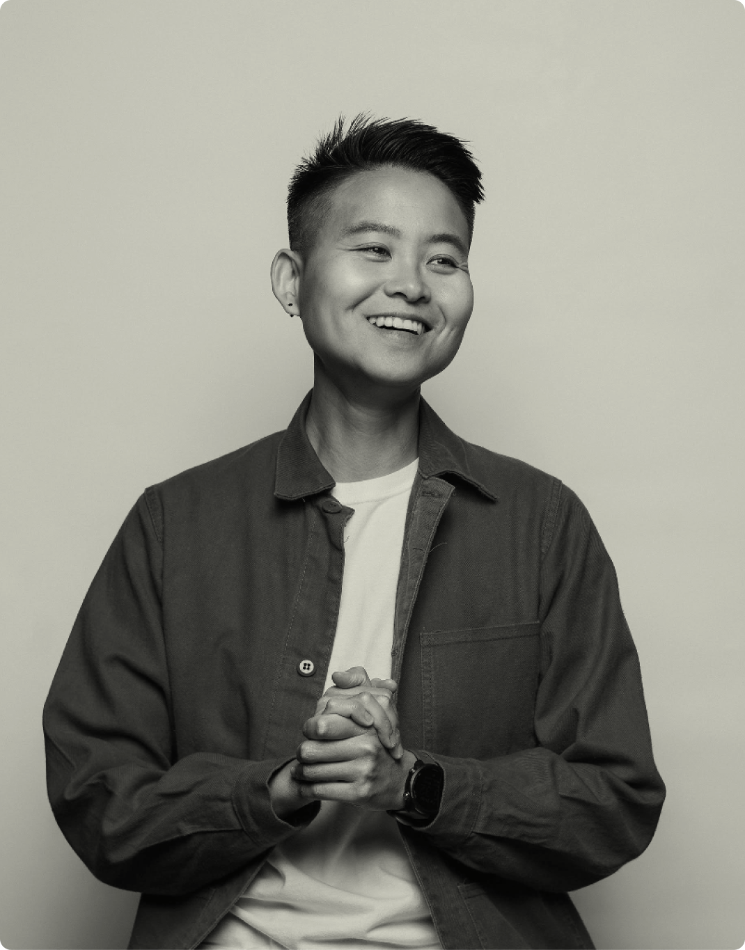 A black and white professional headshot photograph of Katie Chen. She is smiling warmly at the camera. She is wearing an unbuttoned dark overshirt with a white t-shirt underneath, a small earring, and a smartwatch. Her hands are clasped together in front of her chest. Her hair is styled in a modern short cut. The image is shot against a plain light background, creating a clean, professional look.