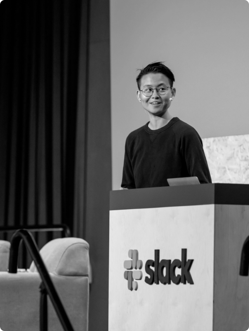 Katie Chen standing at a podium with the Slack logo, delivering a presentation. Katie is wearing glasses and a black sweater, and smiling while looking to the side. The background features a stage setup with curtains and seating, indicating a professional event or conference.