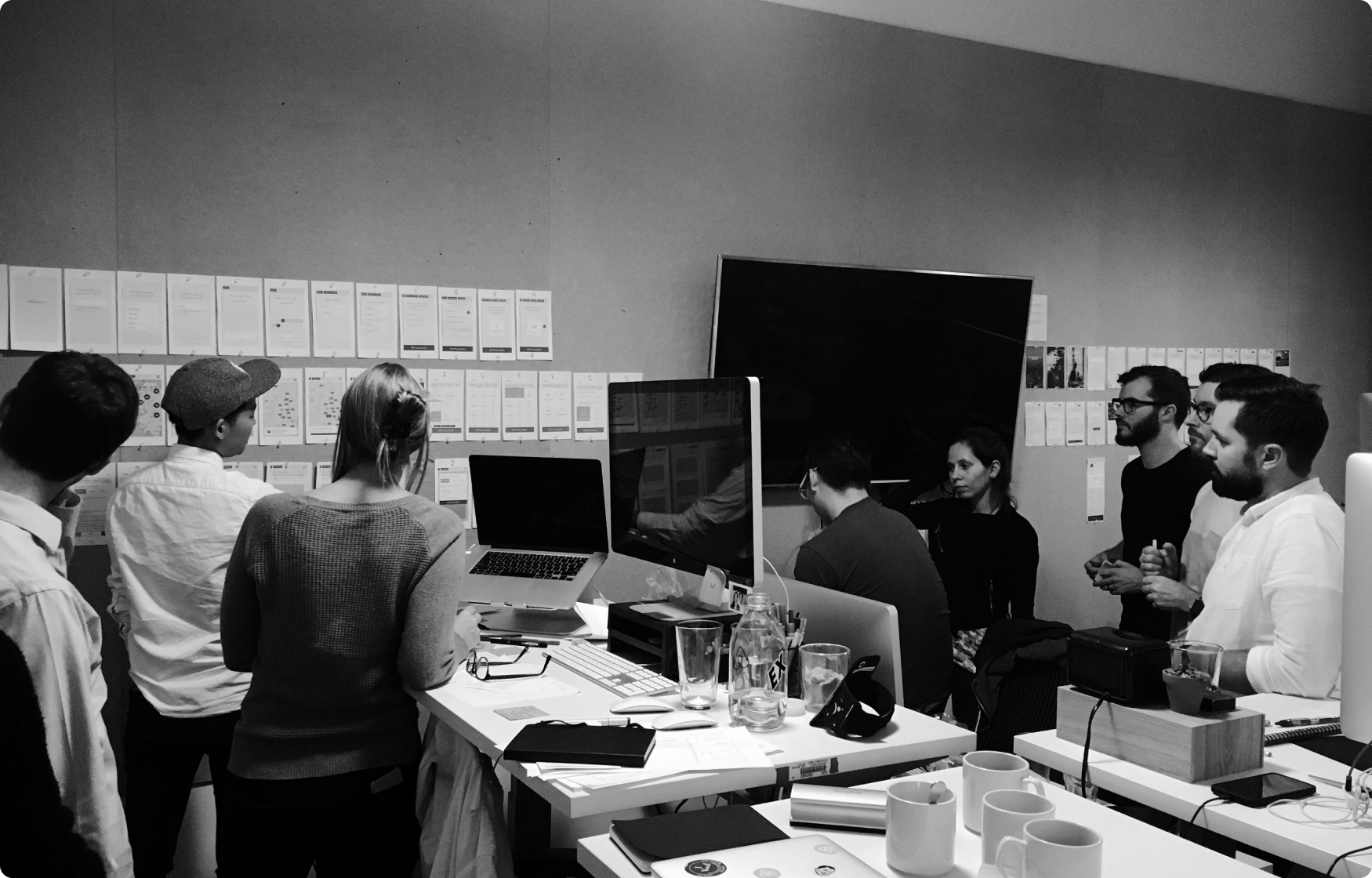 Katie Chen presenting to a team in a design studio. The team is gathered around a workspace with computers and notebooks, attentively looking at a wall covered with design mockups and notes. Katie, wearing a cap, is pointing at the wall while others listen and discuss. The environment is collaborative and focused, with a modern office setup.